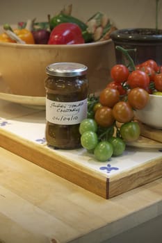 A kitchen setting with a white ceramic bowl of freshly picked small green, red and yellow coloured tomatos cascading from the bowl with a jar of homemade sweet tomato chutney in front with a handwritten label. Set on a wooden kitchen island unit. A bowl of colored peppers is set to the back of the image.