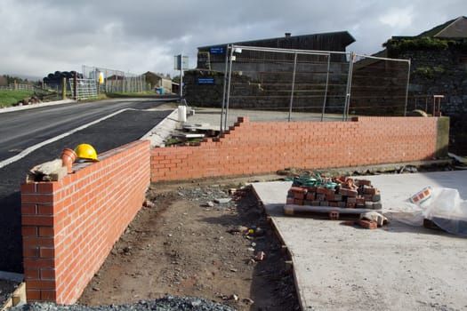 A red brick wall under constructionwith a yellow safety helmet, fencing, a path and a road with buildings.