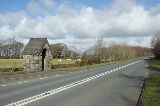 A stone built bus stop next to a tarmac road with lines leading into the distance backed by a cloudy blue sky.