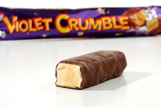 A piece of Voilet Crumble by Nestle chocolate bar in foreground with the wrapper packaging in the background.    Sweet honeycomb coated with milk chocolate.  Focus is to the foreground.