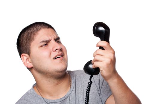 An angry and irritated young man yells into the telephone receiver over a white background.