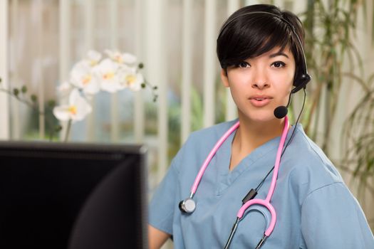 Smiling Attractive Multi-ethnic Young Woman Wearing Headset, Scrubs and Stethoscope Near Her Computer Monitor.