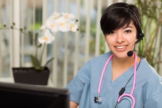 Smiling Attractive Multi-ethnic Young Woman Wearing Headset, Scrubs and Stethoscope Near Her Computer Monitor.