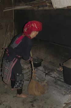 Grandmother of ethnic (minority) Hmong red pompoms. This woman wears the traditional clothing of his tribe and the hair of married women. Before the fireplace she brushes the ashes with the typical brush made of rice straw.