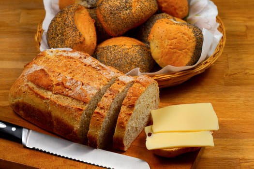 Fresh bread cheese and knife on wooden cutting board with basket with bread rolls in the background