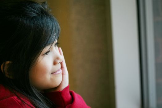 Little Asian girl quietly sitting by window, hand on chin