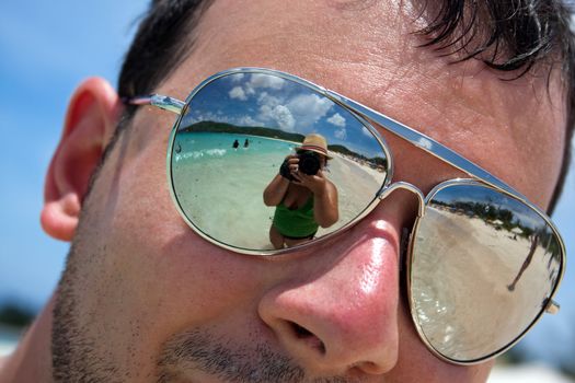 Close up of a man wearing reflective sunglasses in a tropical beach with reflection of the woman photographer in the lens. Shallow depth of field.