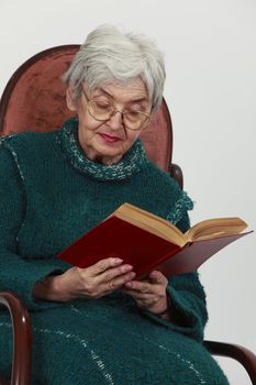 Portrait of an old woman reading a red book against a gray background.