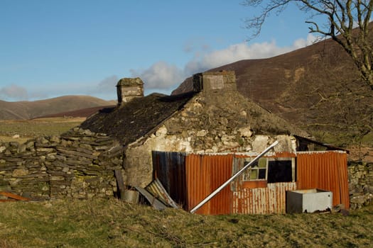 Derelict abandoned building, cottage, with crumbling walls and roof on grass with a metal rusty walled extention.