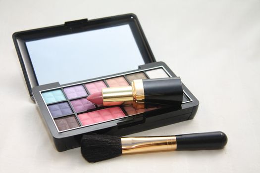 A palet with different shades of eye shadow, pink lipstick and a brush
