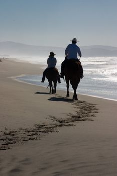 Two riders on horses along the ocean ride away from the camera, backlit by the sun.