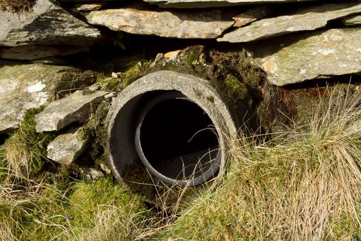 A concrete culvert pipe with plastic inner built into a stone wall with grass and moss.