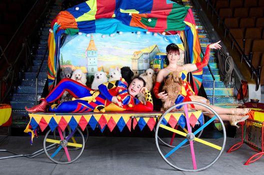 Couple of smiling clowns posing with small dogs on vintage carriage with the painting of the old town