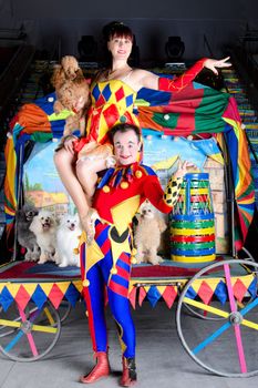 Smiling Harlequin holding Colombina on his shoulders. She carries small red poodle. At the background group of dogs and retro carriage