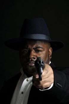 young African American man in a hat and suit with a gun pointed at the camera