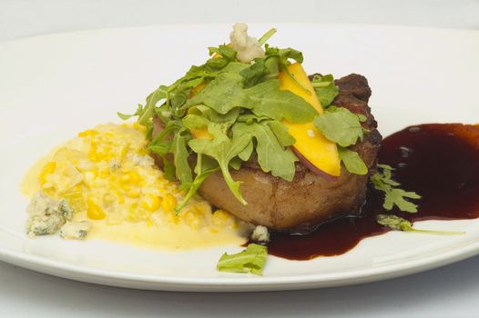 a plate of filt mignon topped with dandelion leaves and peach garnish  with corn and blue cheese and a fruit sauce