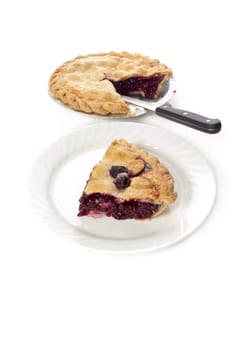 Slice of cherry pie on a white plate sitting on a white background with remainder of pie in pan behind
