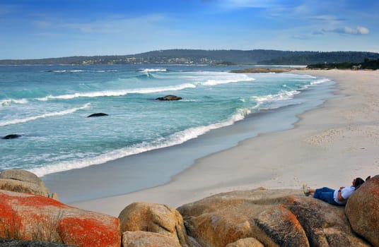 An empty beach in eastern Tasmania, with a solitary person resting on the rocks to the right.