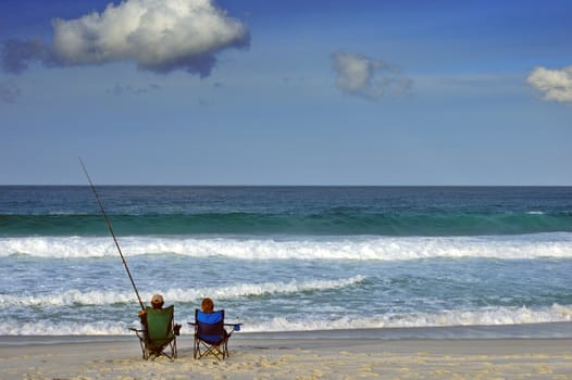 A couple sit on a beach the man fishing, the woman gazing out to sea..