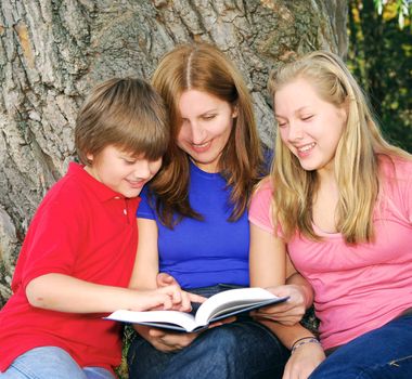 Family of mother and children reading a book under a tree in summer park