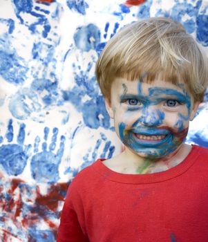 A small girl painted her face and the wall back. The girl is smilling very satisfied and her face is painted with blue, red and green. The wall is painted with these colors also. This picture is a montage.