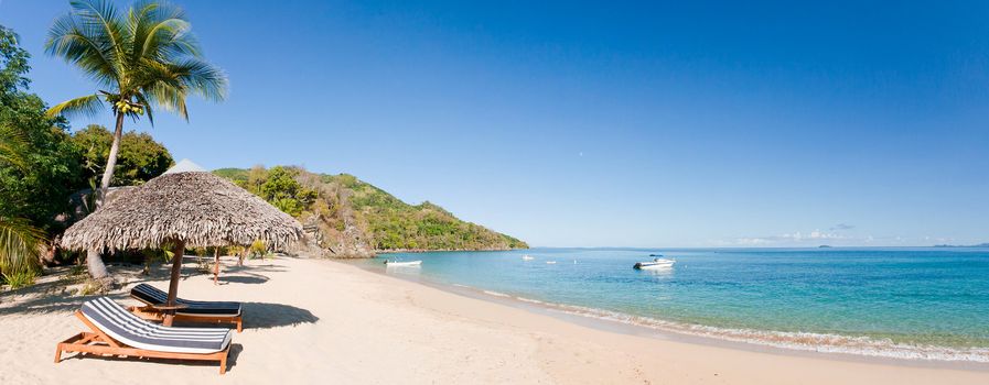 Tropical beach panorama with deckchairs, umbrellas, boats and palm tree