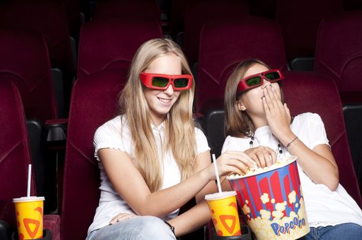 two girls look three-dimensional cinema, sitting in the glasses, eat popcorn, drink drink