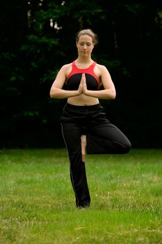 Young adult woman practising yoga on a field surrounded by trees