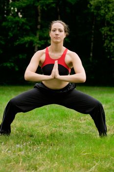 Young adult woman practising yoga on a field surrounded by trees