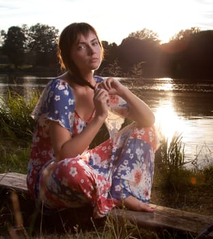 a young woman sitting on a log in front of a lake backlight from the sun lightened up with flash