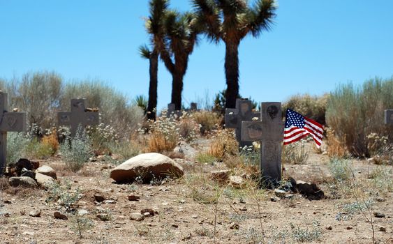 A cross headstone is accented by an American flag in a desert cemetery