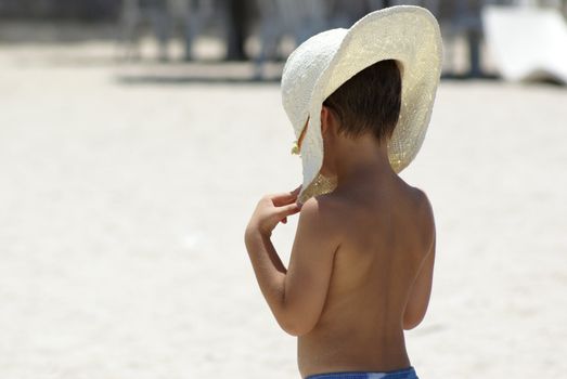 A boy protecting himself with sunshine with his mothers hat at the beach.