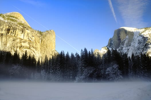 Sunset on the granite peaks in Yosemite National Park with mist rising from the snow cover floor in the valley, an airliner leaving a trail in the sky and snow on top of Half dome.