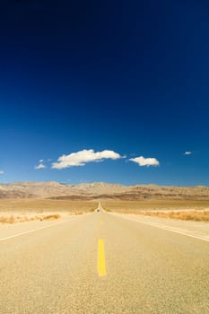 long straight road across Death Valley with a distant motor vehicvle coming towards the camera, a deep blue sky late in the afternoon, and a small puffy clouds in the sky