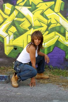 A lovely mature black woman, wearing denim bib overalls with work boots and a white tank top, squats in front of a graffiti-covered block wall.