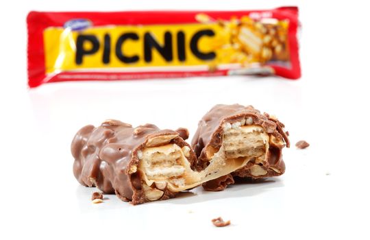 A Cadbury Picnic chocolate bar 46g (1021kj).  Showing wrapper packaging in background and chocolate bar contents in focus in the foreground. Picnic contains nuts, wafer, chocolate, rice crisps and caramel.  Photographed in studio on a white background.