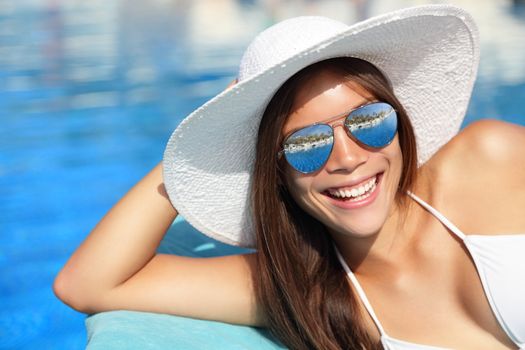 Summer bikini girl smiling by pool. Beautiful young woman wearing beach hat and sunglasses at tropical resort. Beautiful Asian Caucasian female model.