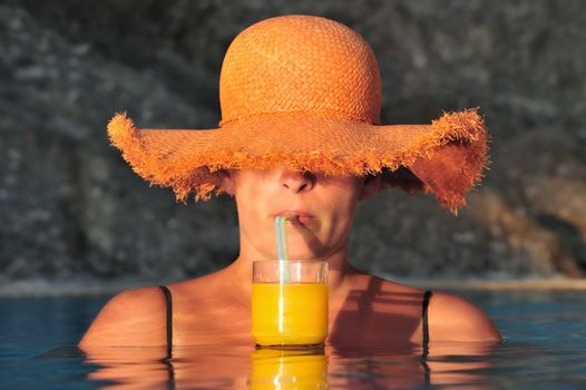 Attractive young woman in straw hat enjoys cool orange juice inside a swimming pool
