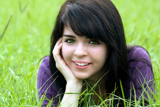 Close-up of a lovely young brunette with a bright, warm smile lying in a field of tall green grass.