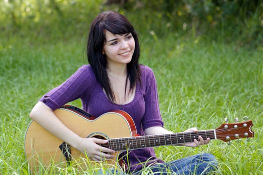 A lovely young brunette sits in a field of tall green grass with her acoustic guitar, looking off camera to frame right.