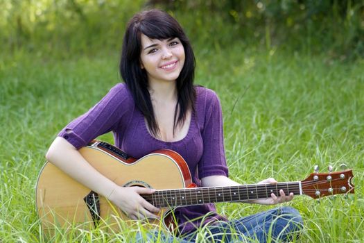 A lovely young brunette sits in a field of tall green grass with her acoustic guitar, looking directly at the camera.