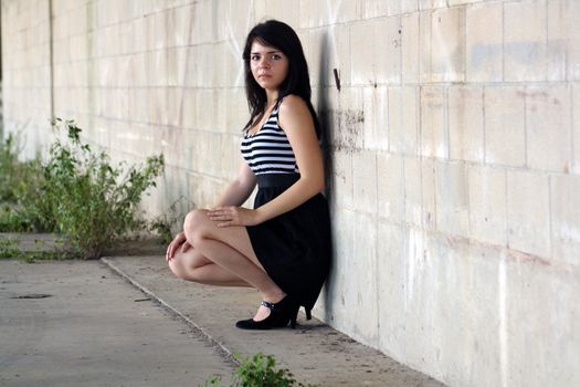 A lovely young brunette squats and leans against a block wall, looking at the camera with a serious facial expression.