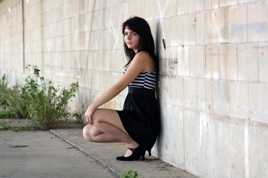 A lovely young brunette squats and leans against a block wall, looking off camera slightly toward frame left with a serious facial expression.