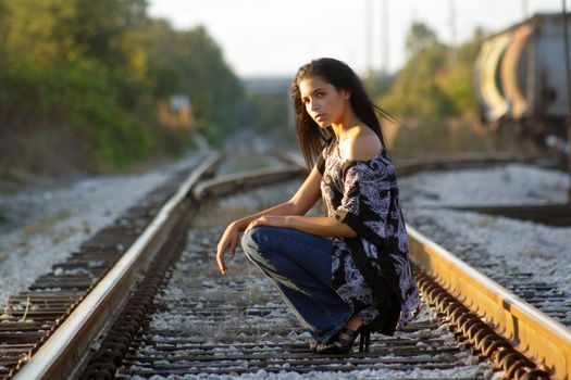 A lovely teenage Latina squats down on a railroad track, looking at the camera.