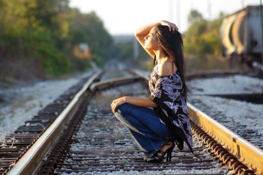 A lovely teenage Latina squats down on a railroad track, facing the sun with her eyes closed.