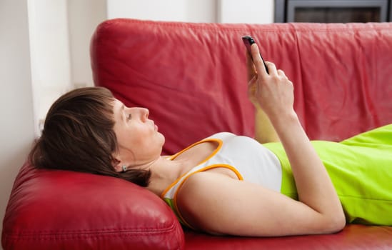Pretty woman lying on red leather sofa and texting with her mobile phone