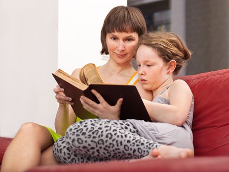 Teenager girl and her mother sitting on sofa and reading book together