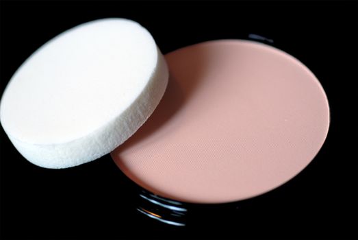 A close-up of a cake of face powder and applicator sponge, on a black background.