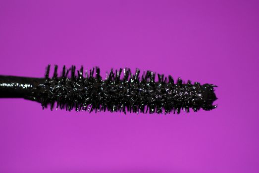 A close-up of the bristles of a mascara wand, with black make-up and a purple background.