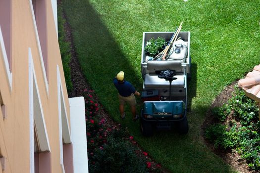 Landscaper and his truck shows a gardner or grounds keeper cart and a man standing in the shade from a hot Florida day while parked on the green grass.
